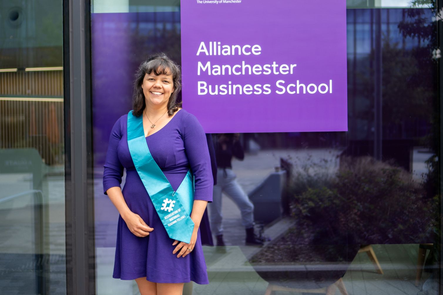 A photo of Jenni Rose wearing a purple dress and has dark brown hair. Jenni is standing outside Manchester Business Alliance School in the photo.